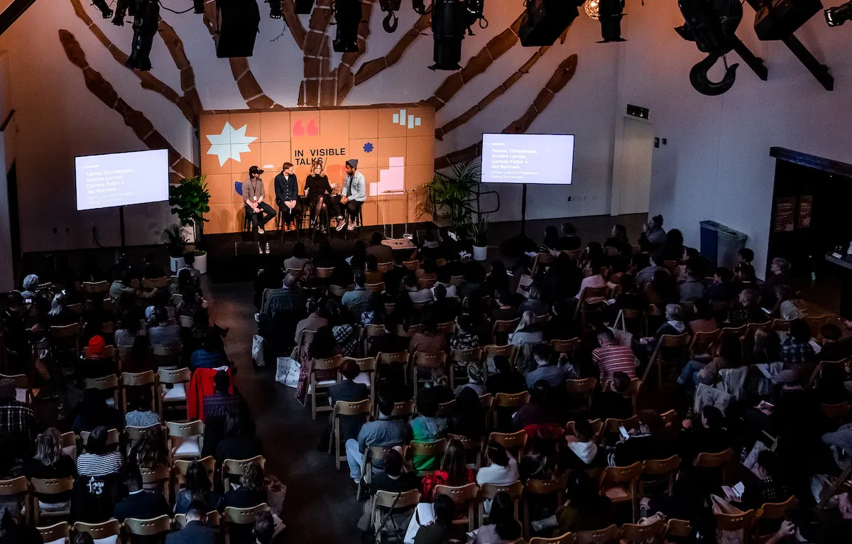 Tanner moderating a panel on stage at In/Visible Talks, with a large audience seated in the room.